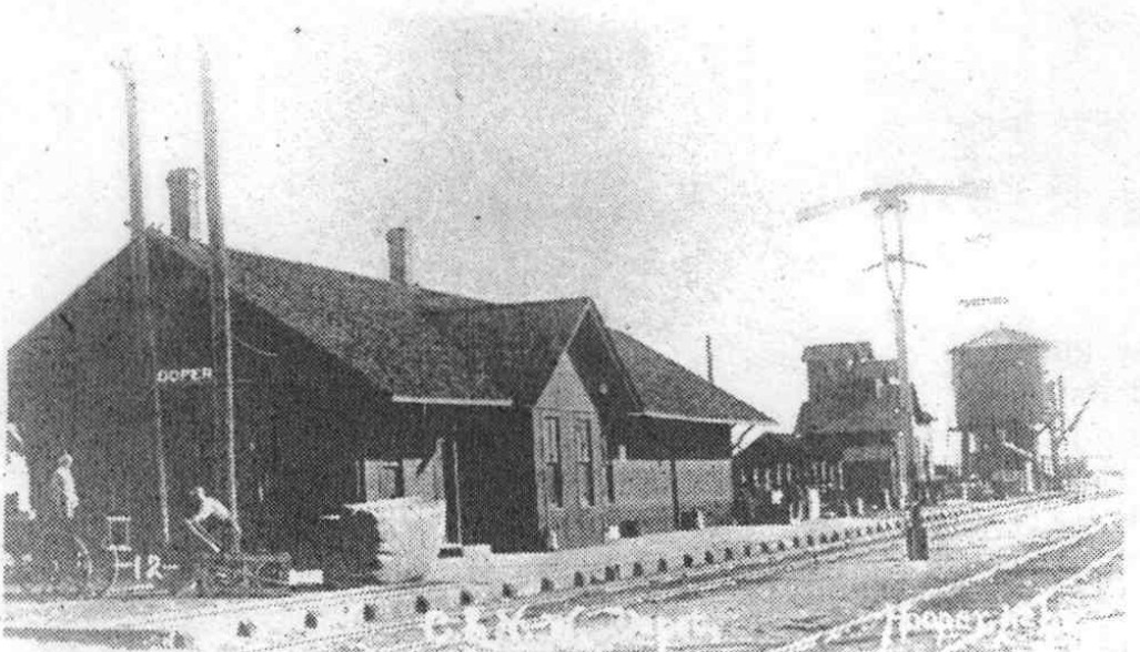 The long platform beside the Hooper depot with stacked materials and railroad water structures beyond.