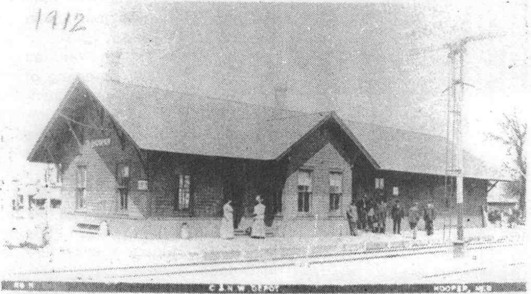 Historic postcard showing the Hooper railroad depot, people beside the platform and tracks in the foreground.